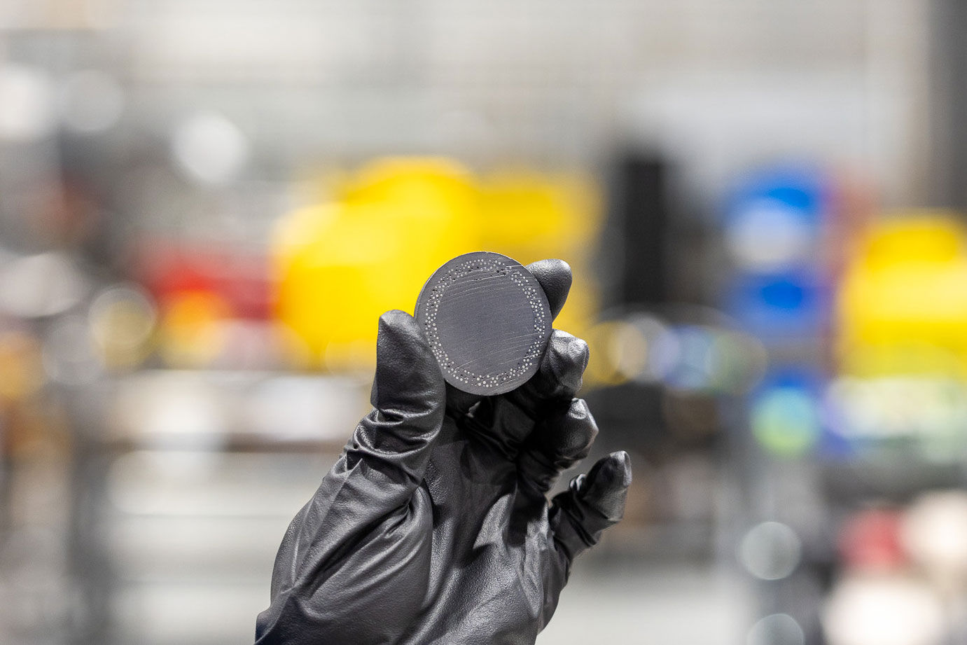 Gloved hand holding a small round metal sample with a patterned surface in a lab setting.