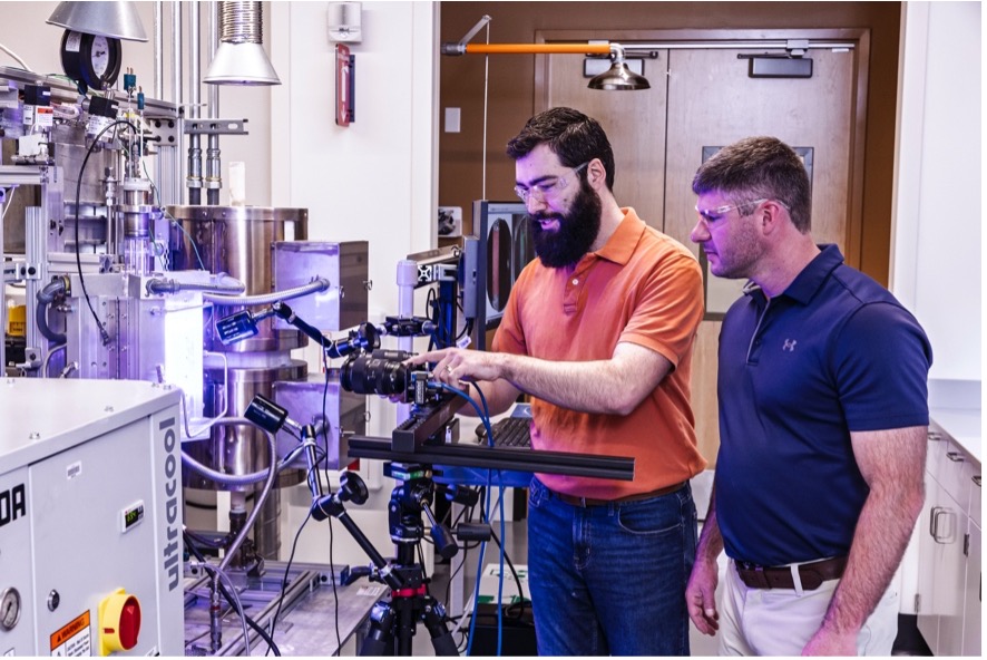 Two researchers adjust a camera system attached to laboratory testing equipment while wearing safety glasses.