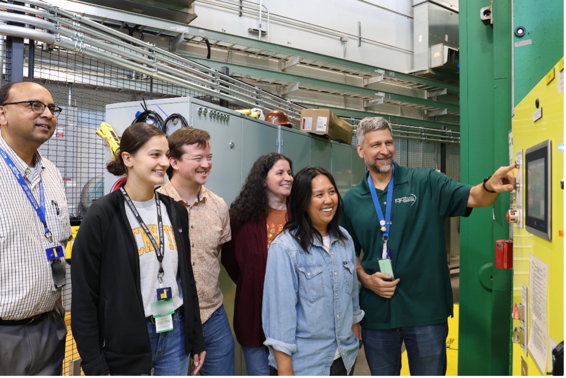 A group of six researchers in a lab stand beside industrial equipment, smiling as one man operates a control panel on a large green machine.