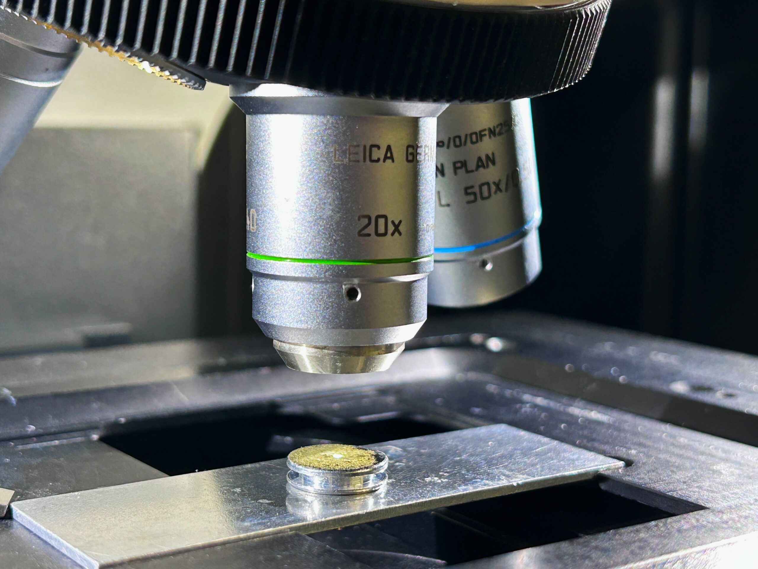 Close-up of a microscope objective lens (labeled 20x) positioned above a glass slide holding a small, circular sample, with metallic stage components visible around it