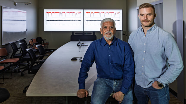 Luiz Leal and Jesse Brown stand in a conference room at Oak Ridge National Laboratory with research data displayed on two monitors behind them. Leal sits on the edge of a conference table while Brown stands beside him during a discussion about nuclear data research.