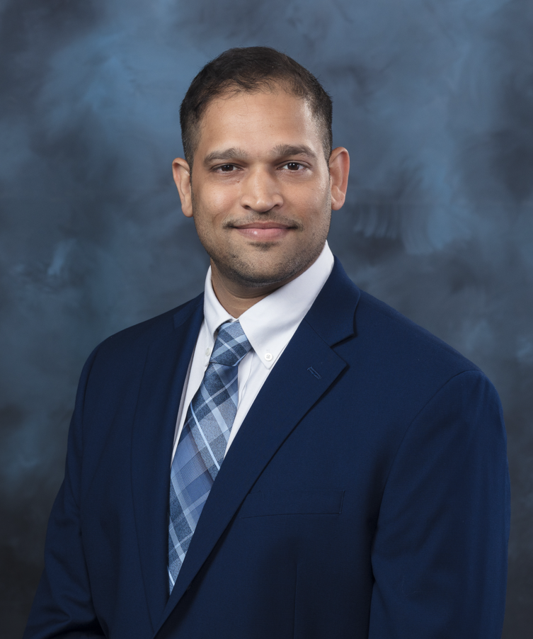 Studio portrait of a man wearing a navy suit and patterned tie, smiling slightly against a blue-gray textured background.