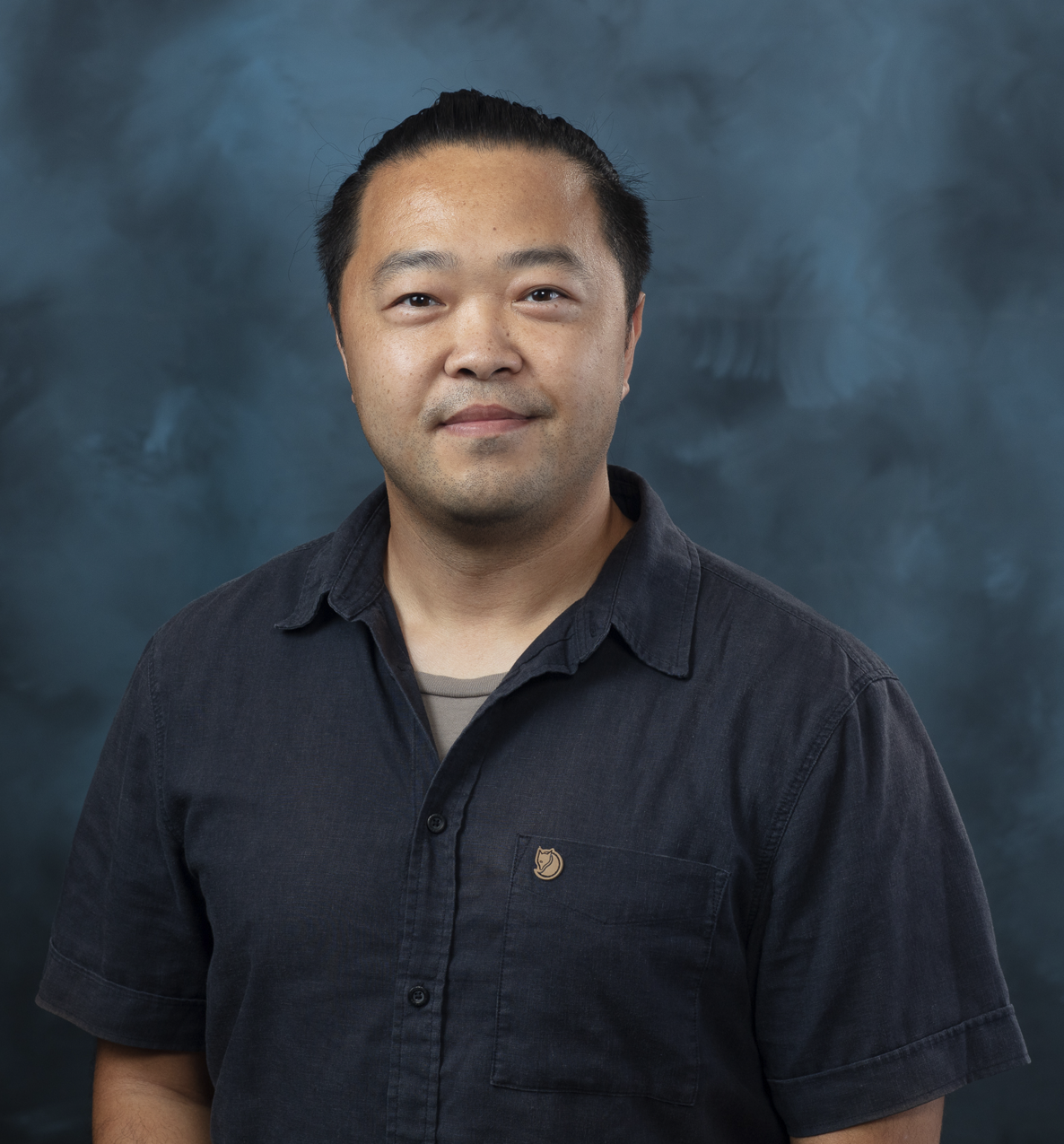 Studio portrait of a man wearing a dark short-sleeve button-up shirt, standing against a blue-gray textured background.