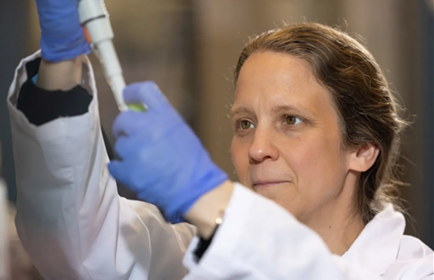 Close-up of a scientist wearing blue gloves and a lab coat, carefully using a pipette to transfer liquid into a small tube.