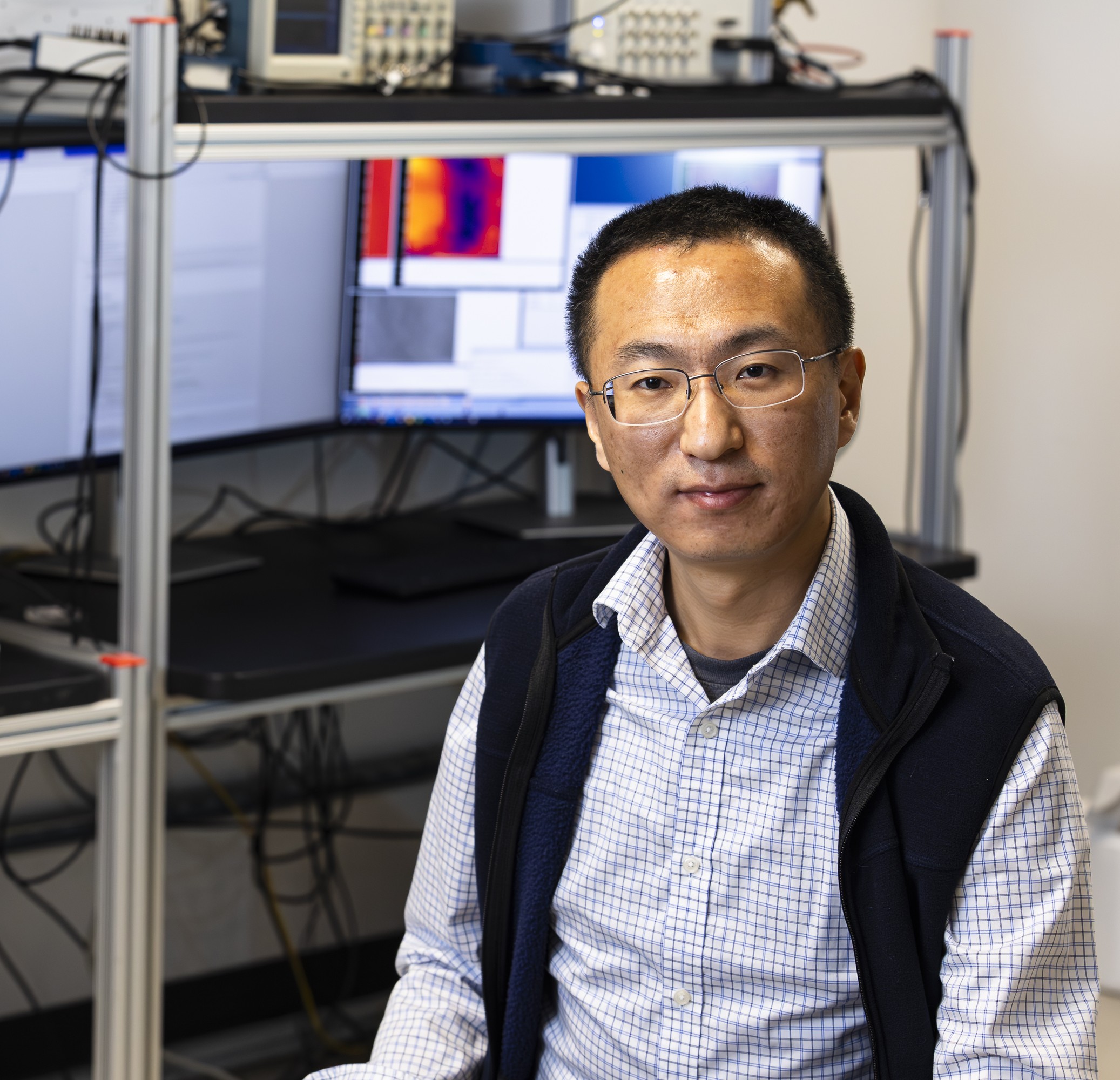 A man in a blue vest and white checked shirt site in front of lab equipment