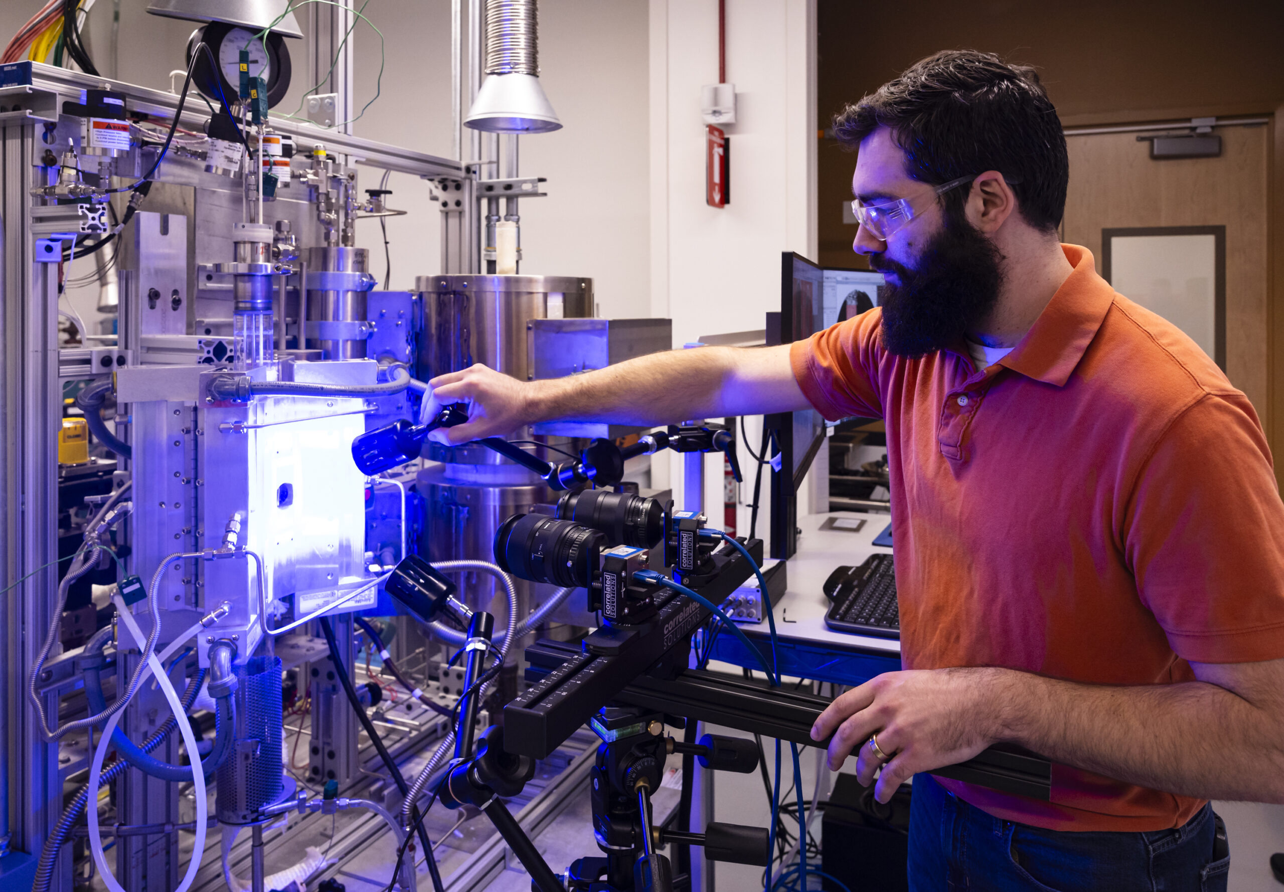 Bearded researcher in safety glasses adjusting a blue-lit laboratory instrument with mounted cameras.