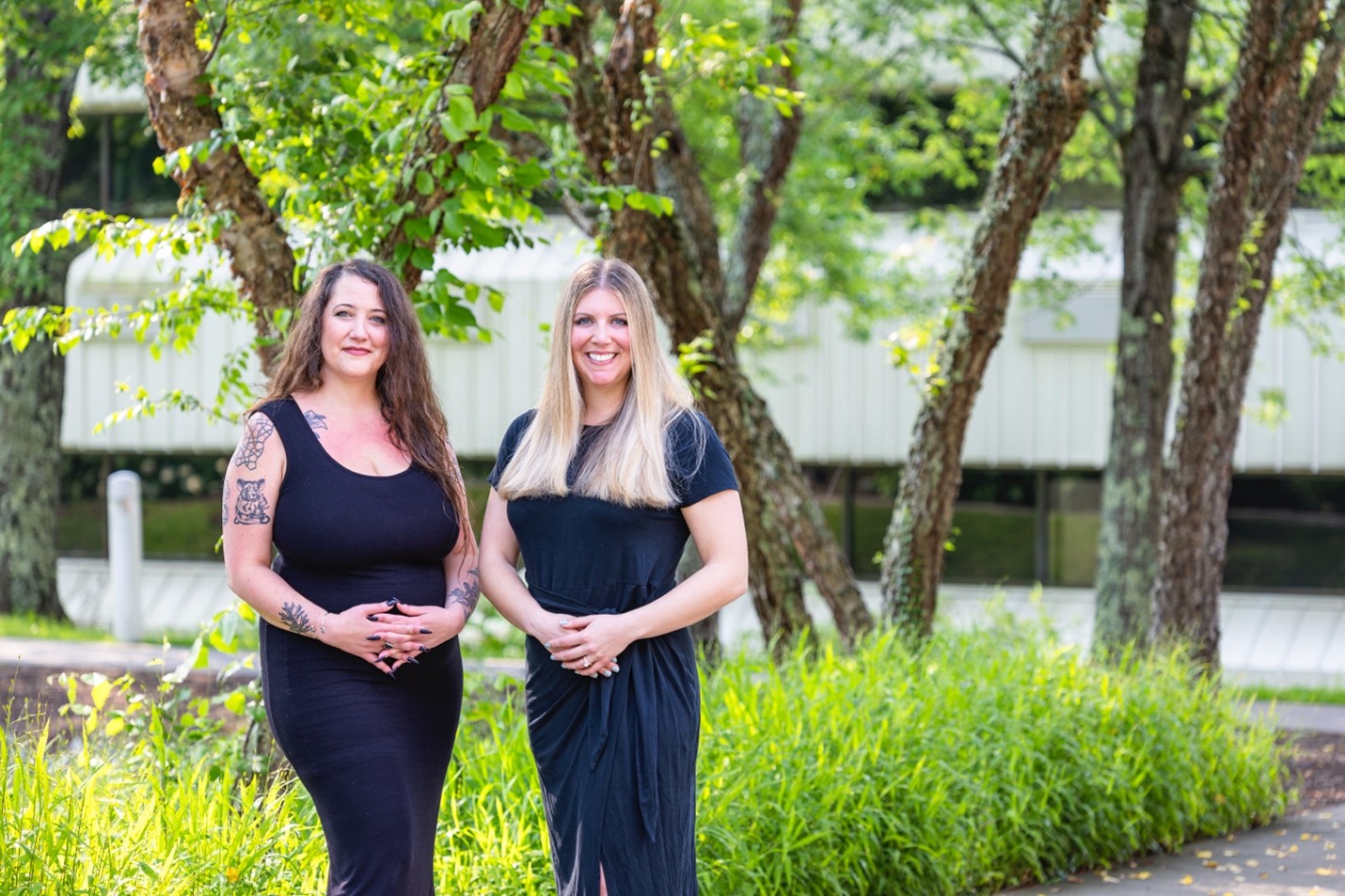 Two women are standing outside in front of trees with black dresses on and their hair down crossing their arms.