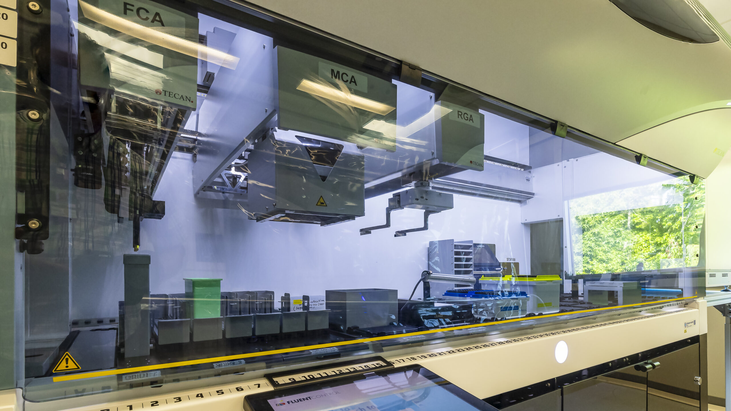 Interior view of an automated laboratory workstation enclosed behind a glass panel, showing multiple labeled modules (FCA, MCA, RGA) mounted on a robotic rail system. Inside are robotic arms, pipetting components, racks, trays, and lab containers arranged along a conveyor-like deck. Reflections of overhead lights appear on the glass, and greenery is visible through a window in the background