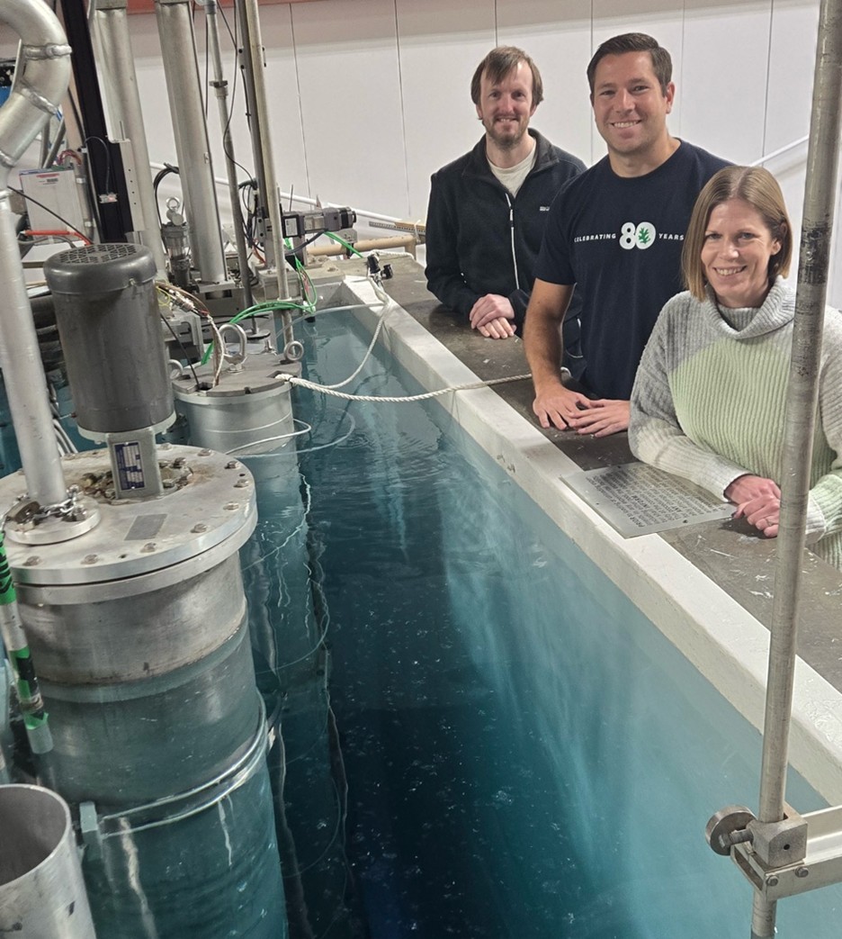 Three researchers stand beside a large laboratory water tank containing a submerged cylindrical metal instrument with attached cables and piping.