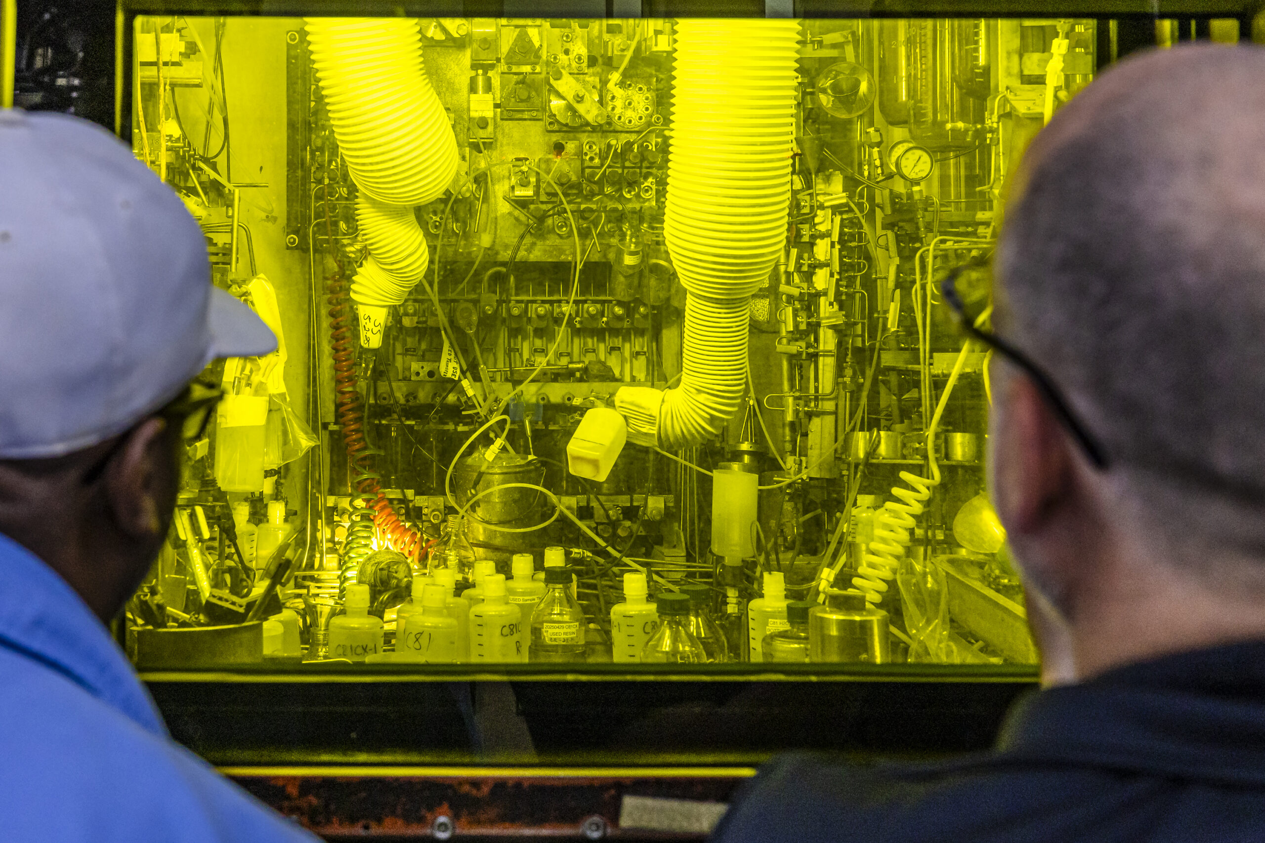 Two people view radiochemical processing equipment through a shielded hot cell window.