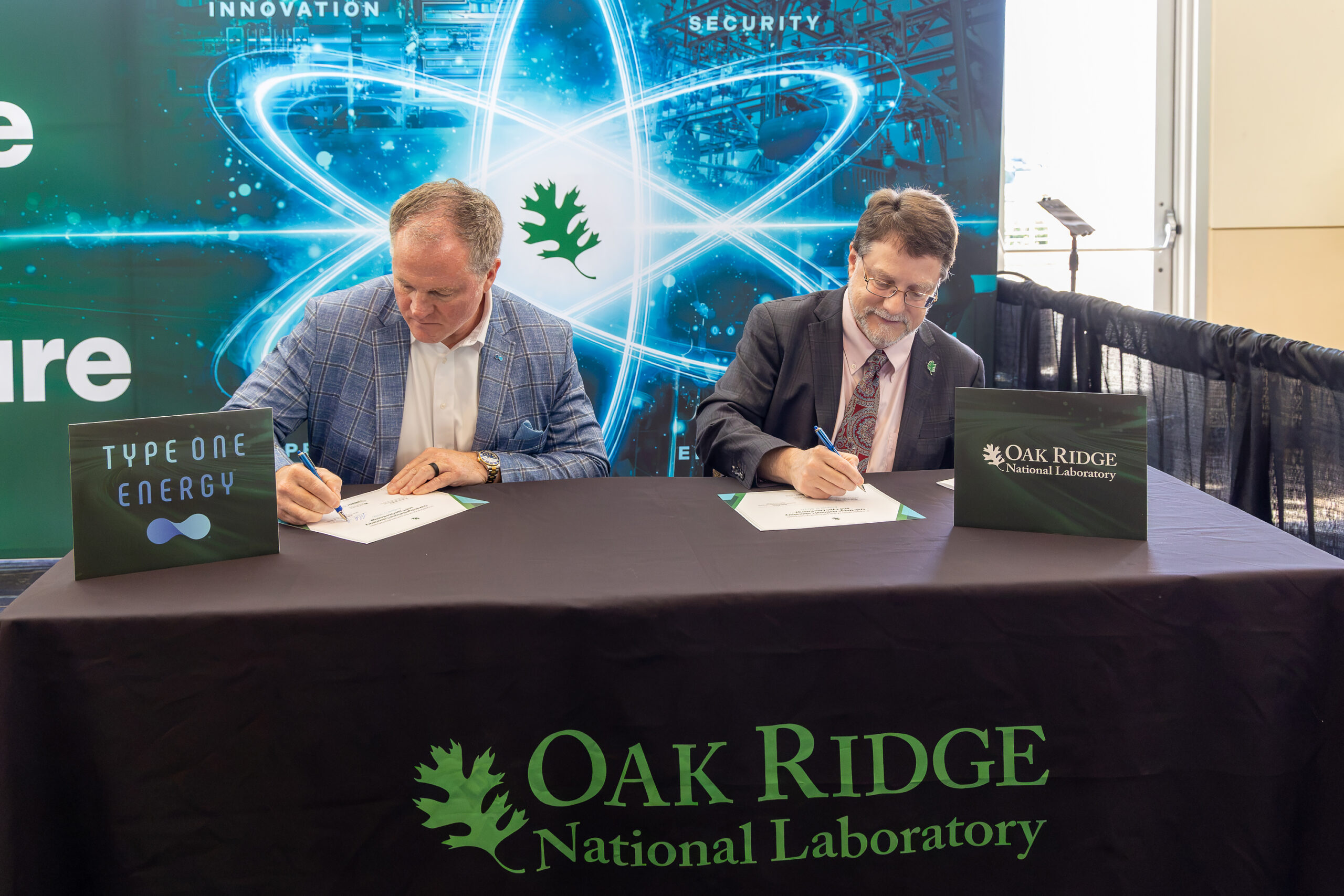 ORNL Director Stephen Streiffer and Mike with Type One Energy sign a piece of paper in front of a nuclear energy symbol and ORNL logo