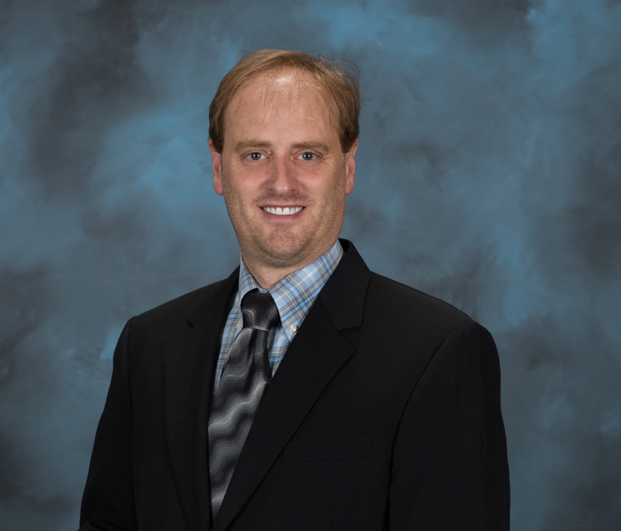 A man in a black suit jacket and tie stands in front of a blue background
