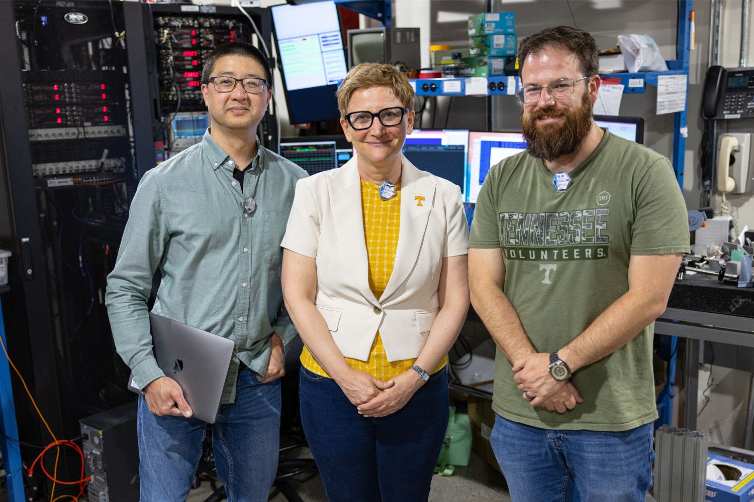 Three people stand together in a laboratory with computer equipment and server racks in the background.