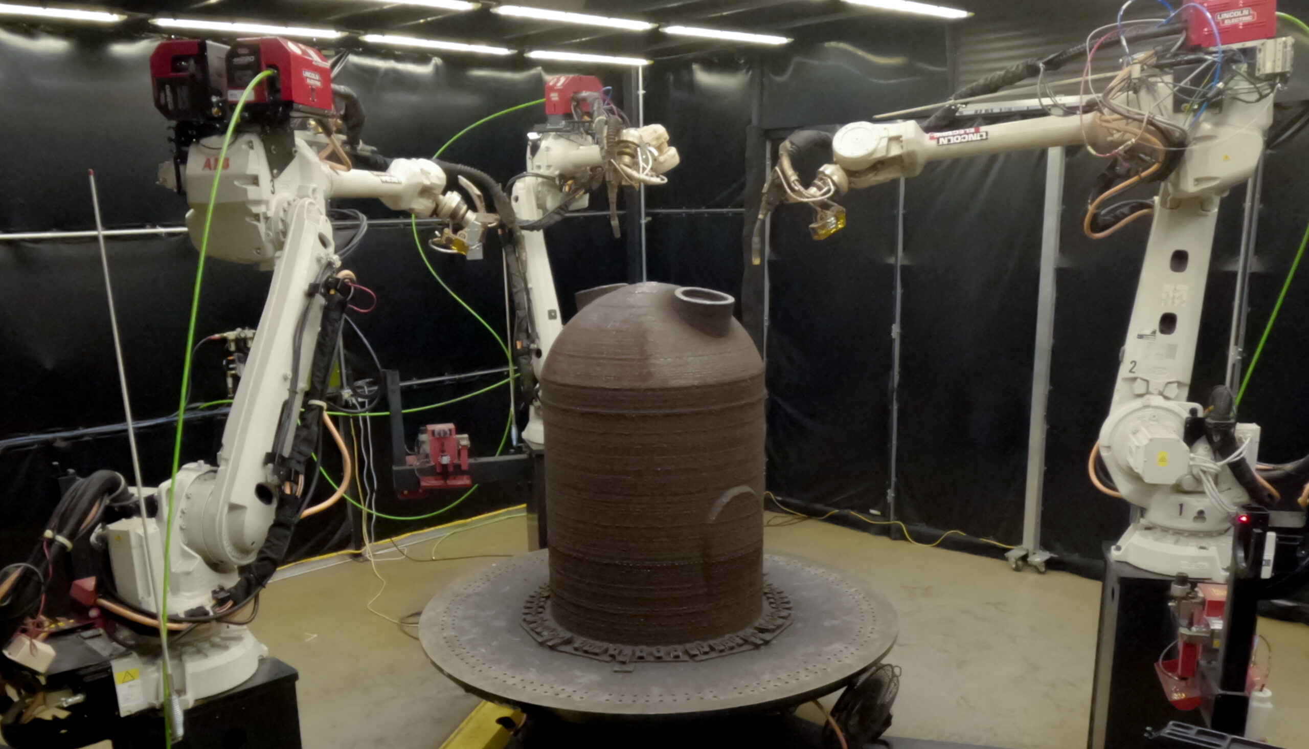 Three robotic arms surround a large 3D-printed metal component.