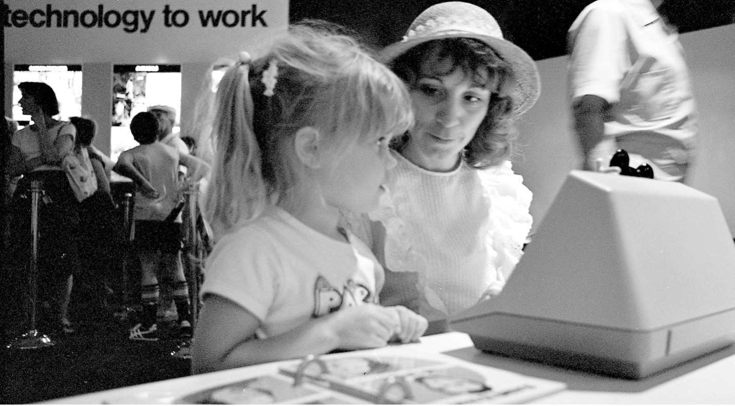 Black-and-white photo of a child and an adult using an early touchscreen exhibit at a technology display, with visitors in the background.