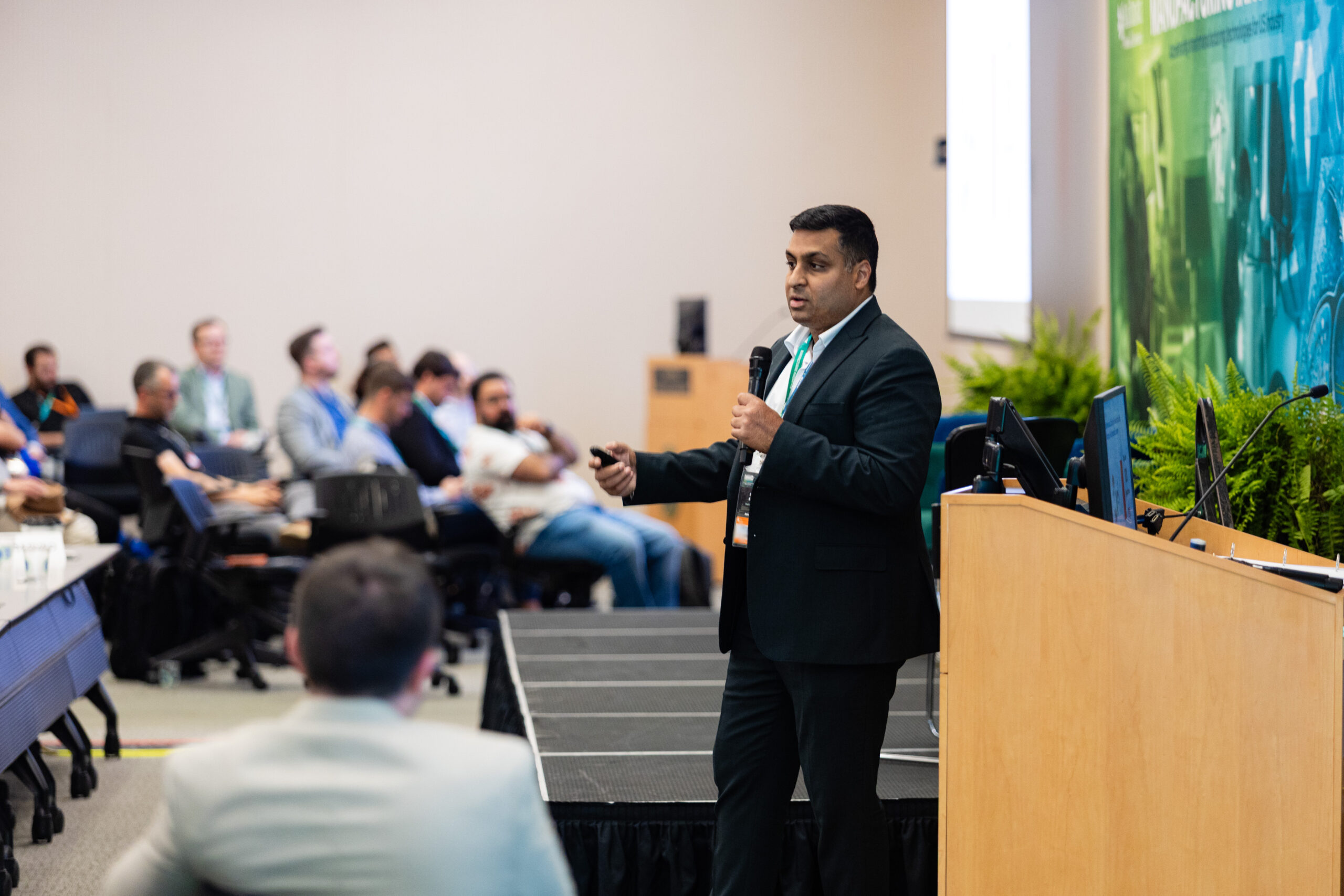 Southeast Asian man in a suit presents in front of a crowd. A wooden podium is in the foreground.