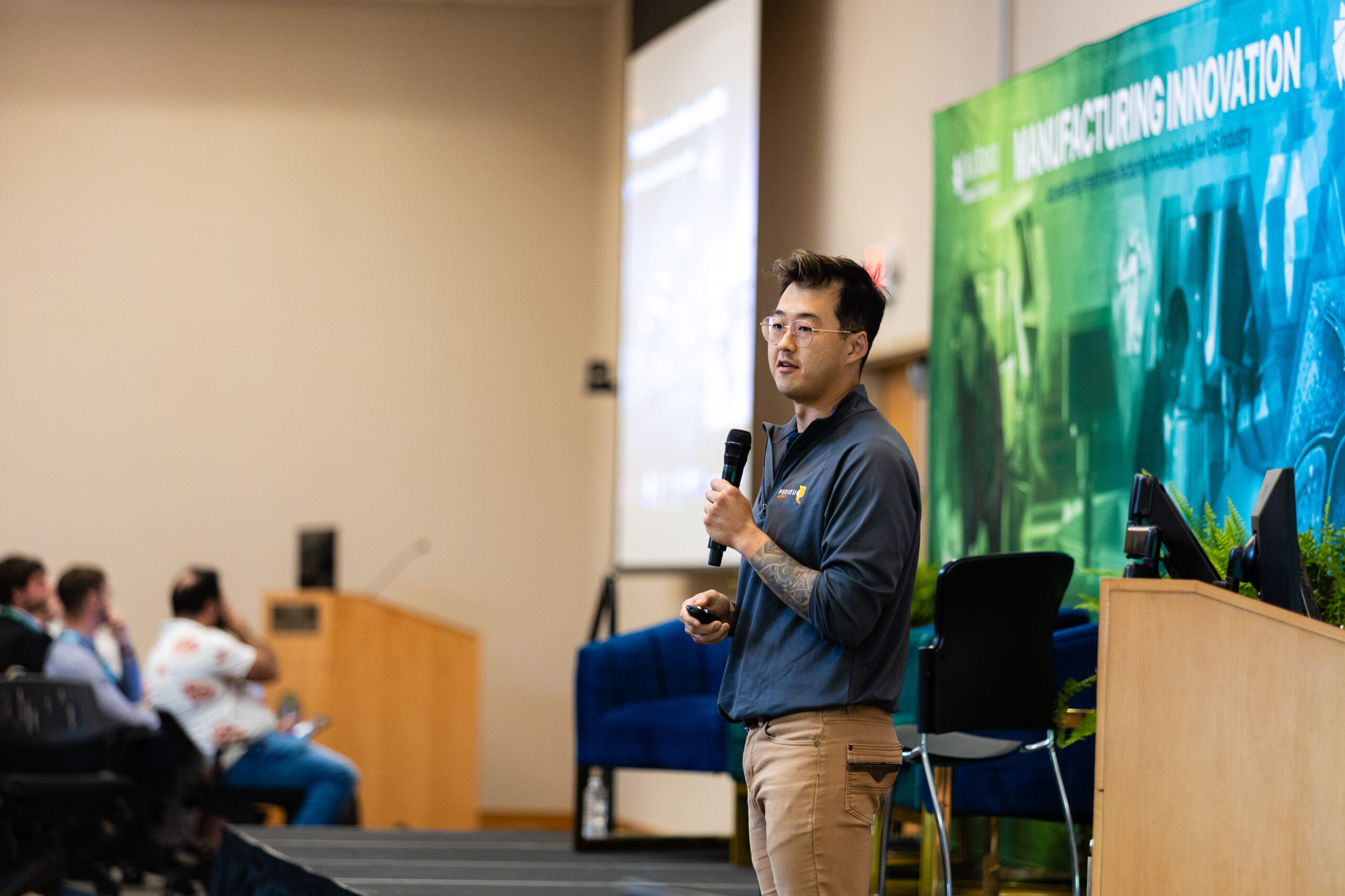Asian American man presents before a conference room. He's wearing a blue shirt and holding a microphone.