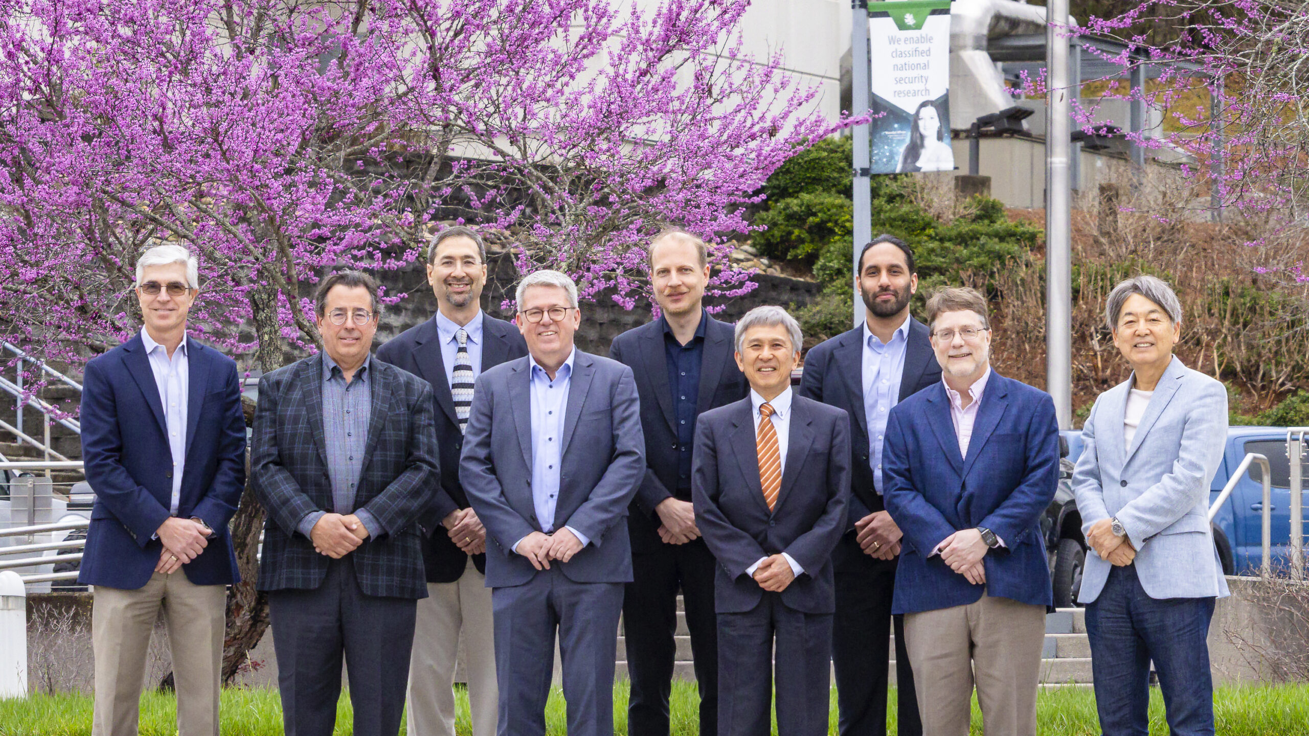 Group photo of nine people standing outdoors in front of blooming pink trees and an Oak Ridge National Laboratory building.