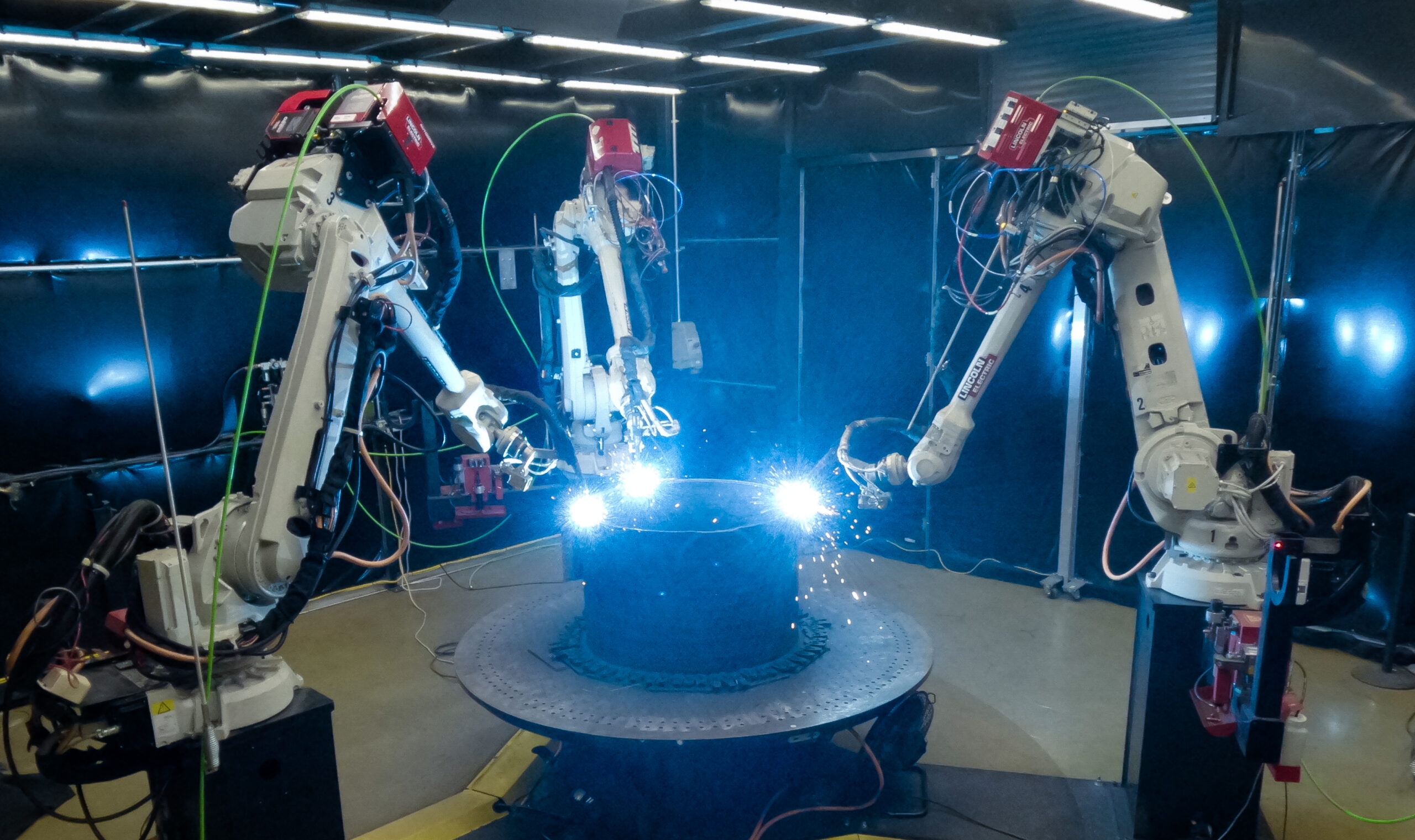 Three robotic arms weld a large metal component.