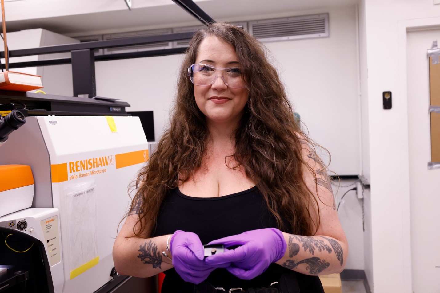 A person wearing safety glasses and purple gloves holds a small sample in a laboratory beside a microscope.
