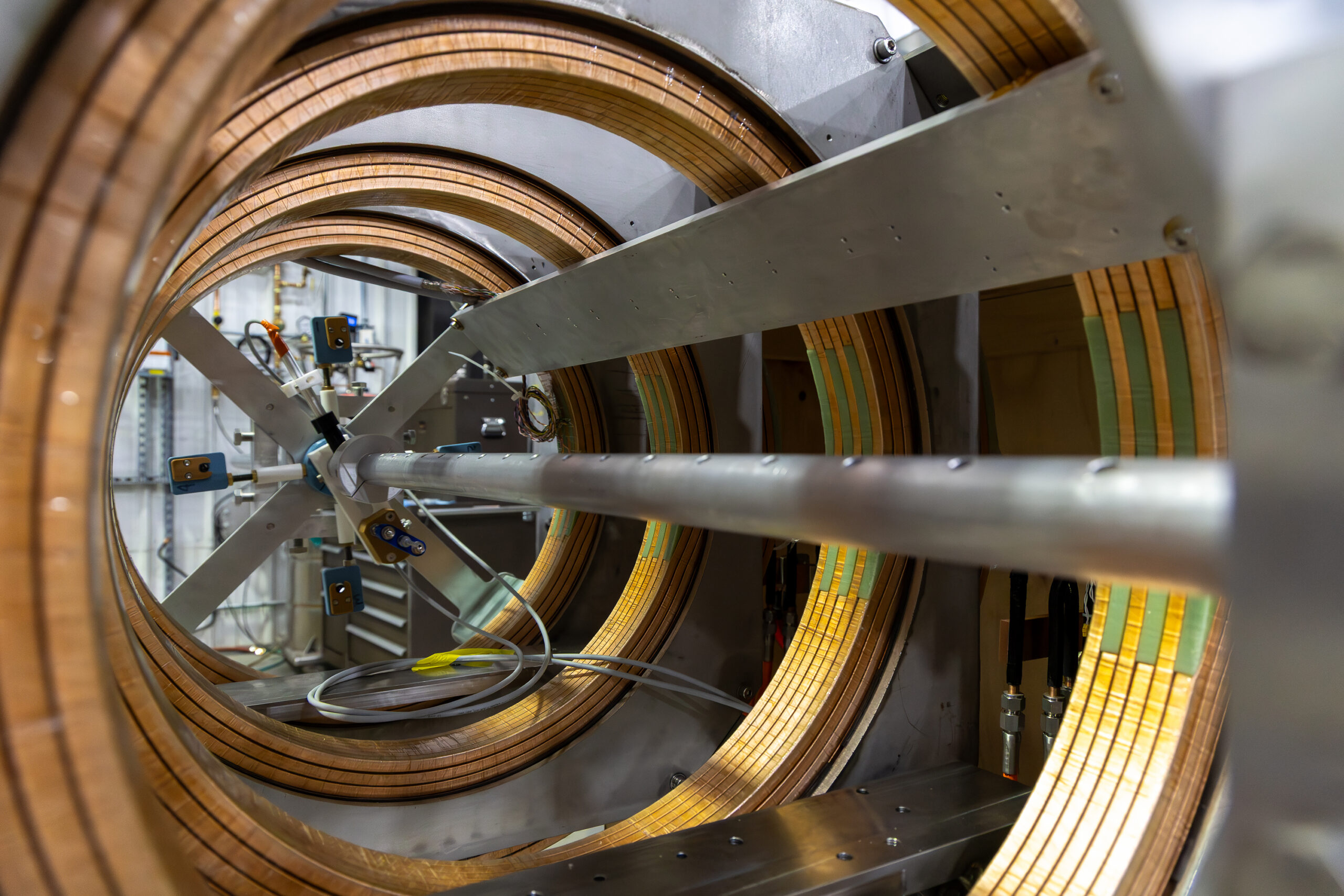 A view looking lengthwise through a cylindrical structure containing multiple concentric copper coils. Electrical cables and instrumentation are attached to the central support rod, with laboratory equipment visible through gaps in the coil assembly.