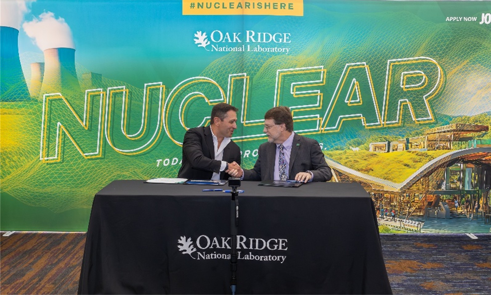 Two men in business attire sit at a table covered with an Oak Ridge National Laboratory cloth, shaking hands during a signing event. Behind them is a large green and yellow banner that reads “NUCLEAR” with images of cooling towers and stylized energy graphics.